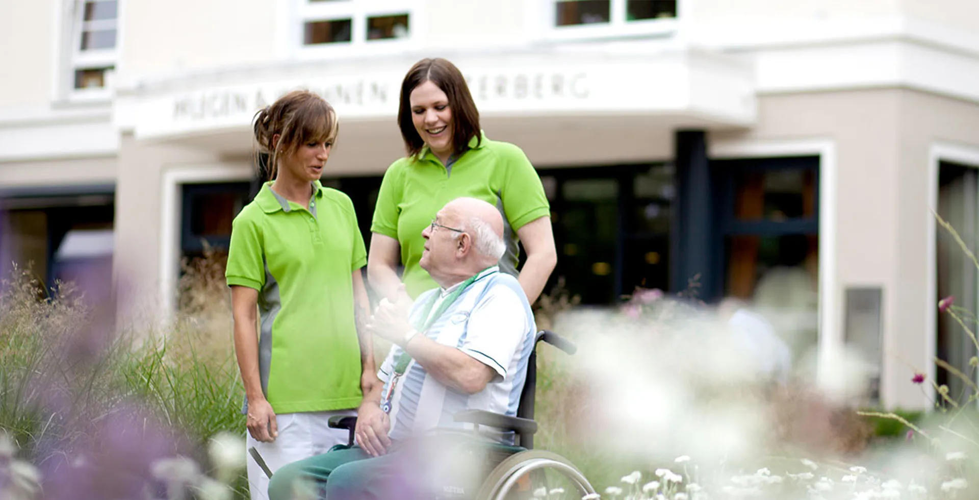 Zwei Pflegekräfte begleiten einen Senioren im Rollstuhl durch den Garten von PFLEGEN & WOHNEN ALSTERBERG.