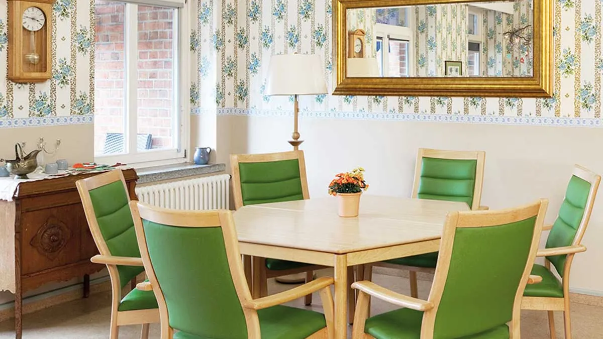 Tisch mit sechs grünen Stühlen in einem Zimmer im PFLEGEN & WOHNEN HAMBURG in Altona. An der Wand hängen ein Spiegel und eine Pendeluhr über einer Kommode.