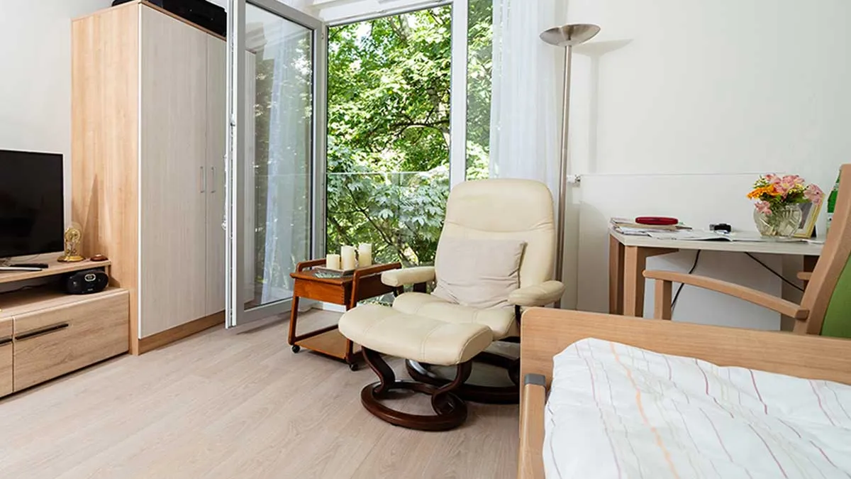 Zimmer im PFLEGEN & WOHNEN HAMBURG-Standort Marienthal mit Sessel, Bett, Schreibtisch mit Stuhl, Schrank und einem Fernseher.