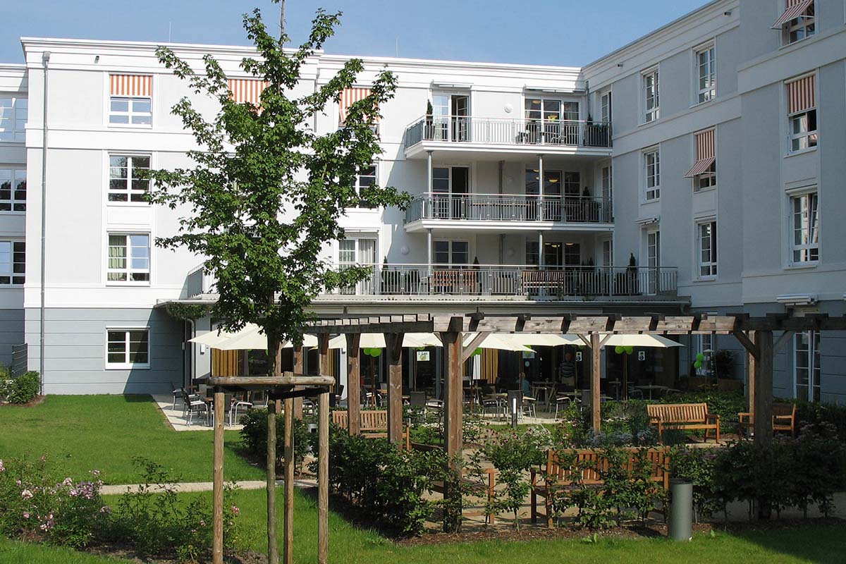 Außenansicht von PFLEGEN & WOHNEN HAMBURG Holstenhof mit Gartenbereich und überdachter Terrasse mit Sitzmöglichkeiten.